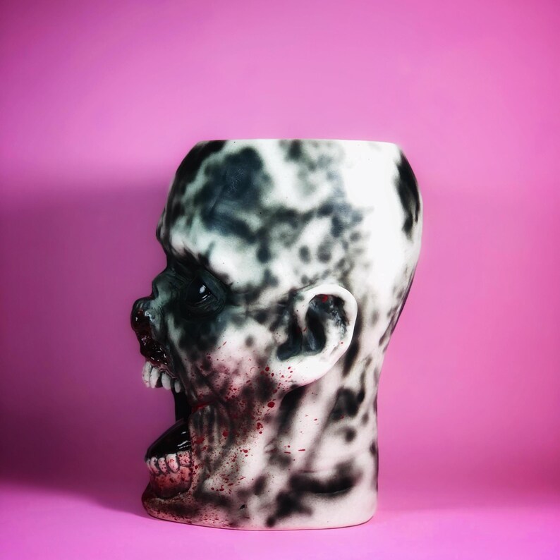 Może przedstawiać: Biała ceramiczna doniczka w kształcie głowy zombie z czarnymi i czerwonymi detalami malowanymi. Gęba zombie jest otwarta i widać zęby. Doniczka znajduje się na r&oacute;żowym tle.