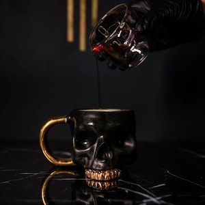 May include: A black skull-shaped mug with a gold handle and gold teeth is being filled with a red liquid from a clear glass container.