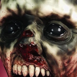 Może przedstawiać: Zbliżenie na realistyczną maskę zombie z krwawą, otwartą paszczą i odsłoniętymi zębami. Maska ma szarą i zieloną karnację z realistycznymi szczeg&oacute;łami, w tym zmarszczkami i rozpryskami krwi.
