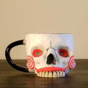 Puede incluir: Taza de cerámica blanca con forma de calavera y asa negra. La taza presenta mejillas y cuencas de los ojos rojas en espiral, una boca roja y una hilera de dientes. Una adición única y espeluznante a cualquier cocina.