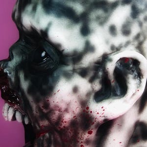 Może przedstawiać: Zbliżenie na realistyczną maskę zombie z szczeg&oacute;łową, groteskową twarzą. Maska jest pomalowana czarnymi i białymi wzorami w stylu rozprysku oraz czerwonymi plamami krwi. Maska ma realistyczne ucho i otwarte usta z zębami.