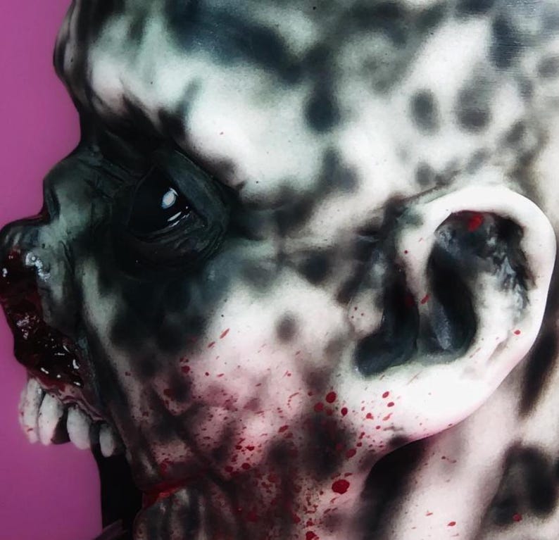 Może przedstawiać: Zbliżenie na realistyczną maskę zombie z szczeg&oacute;łową, groteskową twarzą. Maska jest pomalowana czarnymi i białymi wzorami w stylu rozprysku oraz czerwonymi plamami krwi. Maska ma realistyczne ucho i otwarte usta z zębami.