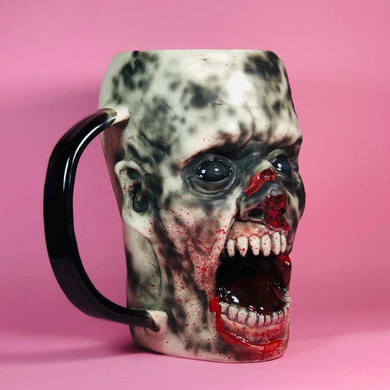 Może przedstawiać: Ceramiczny kubek w kształcie głowy zombie z czarnym uchwytem. Kubek jest pomalowany realistycznymi detalami, w tym krwią, zębami i otwartą paszczą.