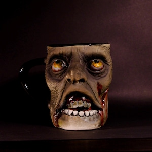 Puede incluir: Una taza de café de cerámica con forma de cabeza de zombi con asa negra. El zombi tiene ojos amarillos, una boca ensangrentada y una tez verde.
