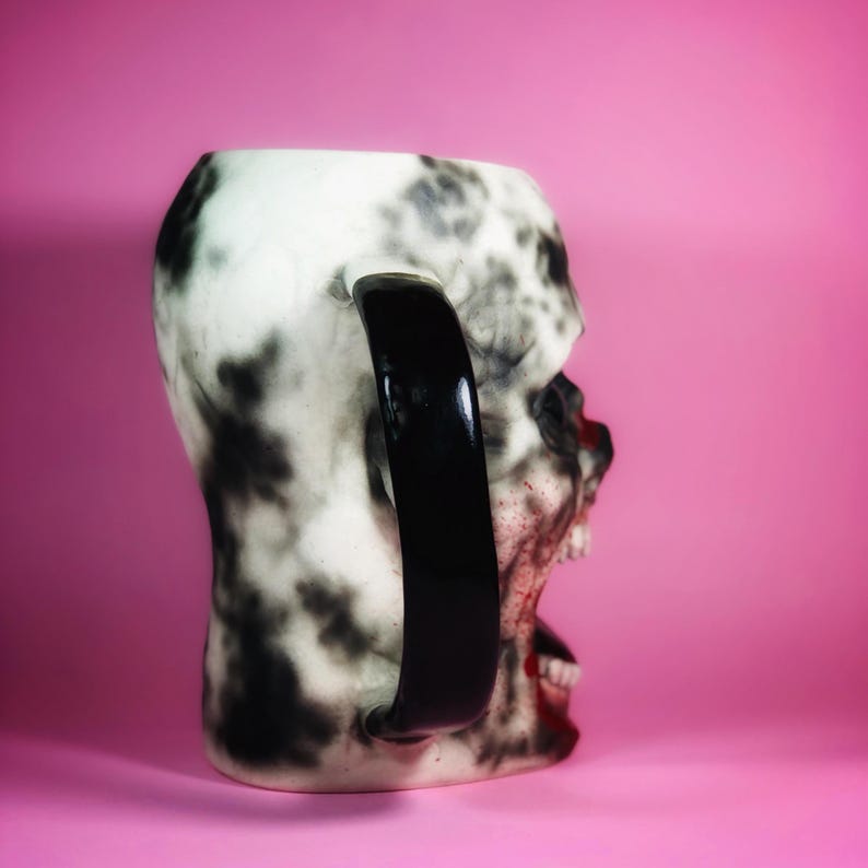 Może przedstawiać: Biały ceramiczny kubek w kształcie czaszki z czarno-białym wzorem w stylu rozbryzgu. Kubek ma czarny uchwyt i pomalowane na czerwono usta.
