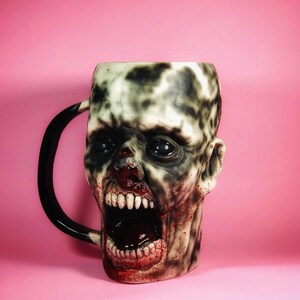 Może przedstawiać: Ceramiczny kubek w kształcie głowy zombie z szeroko otwartymi ustami, pokazujący zęby i krew. Kubek jest biały z czarnymi i szarymi detalami, a oczy zombie są czarne.