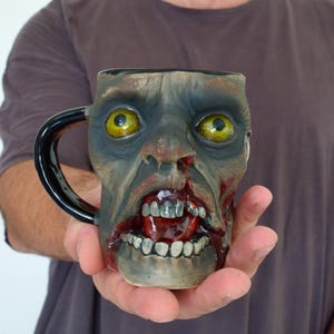 Pode incluir: Uma caneca de café de cerâmica em forma de cabeça de zumbi com uma alça preta. O zumbi tem um rosto verde com sangue vermelho pingando de sua boca e olhos amarelos.