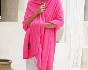 May include: A bright hot pink knit shawl draped over the shoulders, featuring a soft texture and ribbed edges. The shawl is worn over plain white trousers, creating a vibrant contrast. The garment is long and rectangular, providing full coverage.