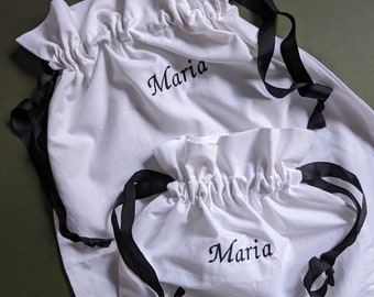 May include: Two white fabric drawstring bags with black ribbon ties. The larger bag has the name "Maria" embroidered on it. The smaller bag also has the name "Maria" embroidered on it.