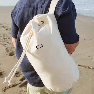 Puede incluir: Una bolsa de lona blanca con un monograma bordado en azul marino "C.J." La bolsa se lleva como mochila por una persona en una playa.