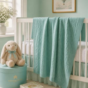 May include: A soft, light teal cable-knit blanket draped over a white wooden crib. The blanket features a vertical braided pattern with a ribbed border. The crib is styled with a light teal bed skirt and a plush cream-colored rabbit toy.