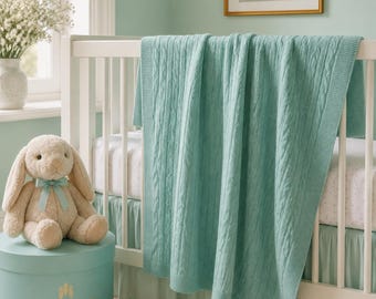 Può includere: Una morbida coperta per neonati in maglia a trecce color verde chiaro drappeggiata su una culla bianca. La coperta presenta un classico motivo a trecce verticali con bordo a coste. La culla è abbinata a un coniglio di peluche beige e una scatola.