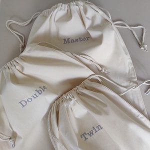 Op de afbeelding: Drie off-white katoenen zakjes met trekkoord in verschillende maten. Elk zakje is voorzien van een grijs geborduurd label. De grootste tas is gelabeld Master, de middelste tas Double en de kleinste tas Twin. Elk zakje heeft een wit koord.