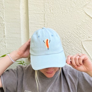 May include: Light blue baseball cap with an embroidered cartoon hot dog design. The hot dog has a smiling face and outstretched arms. The cap is a classic six-panel design with a curved brim.