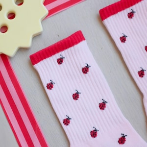 May include: A pair of light pink ribbed socks featuring a repeating pattern of small red ladybugs with black spots. The socks have a solid red cuff at the top and a red heel section. A large, pale yellow, star-shaped button is visible nearby.