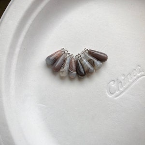 Peut inclure: Un ensemble de huit pendentifs en agate en forme de goutte avec une b&eacute;li&egrave;re en argent. L'agate est un m&eacute;lange de brun, blanc et gris avec un motif tourbillonnant.