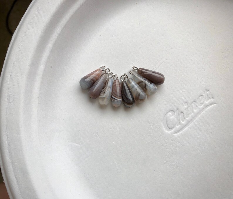 Peut inclure: Un ensemble de huit pendentifs en agate en forme de goutte avec une b&eacute;li&egrave;re en argent. L'agate est un m&eacute;lange de brun, blanc et gris avec un motif tourbillonnant.