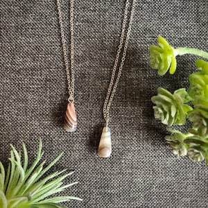 Peut inclure: Deux colliers en argent avec des pendentifs en agate. Les pierres sont de couleur brun clair et gris avec un motif tourbillonnant. Les colliers sont sur un fond de tissu gris.