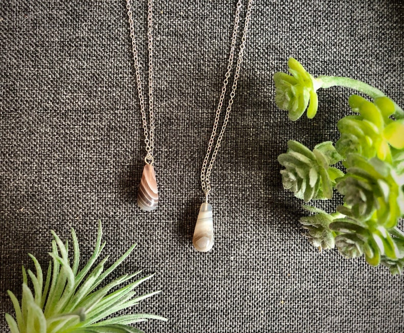 Peut inclure: Deux colliers en argent avec des pendentifs en agate. Les pierres sont de couleur brun clair et gris avec un motif tourbillonnant. Les colliers sont sur un fond de tissu gris.