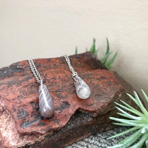 Peut inclure: Deux colliers en argent avec des pendentifs en agate en forme de larme. Les pendentifs sont gris clair avec des rayures blanches et noires. Les colliers sont pr&eacute;sent&eacute;s sur une roche brune.