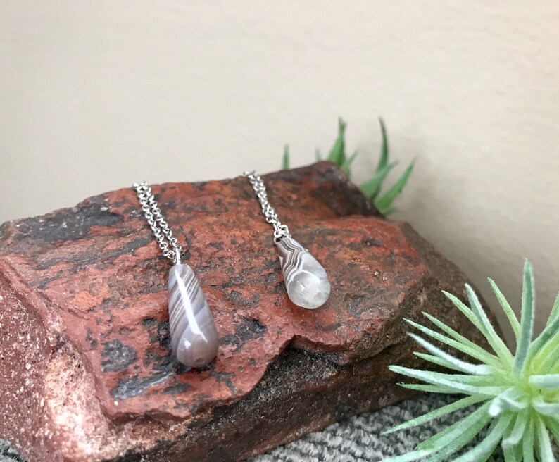 Peut inclure: Deux colliers en argent avec des pendentifs en agate en forme de larme. Les pendentifs sont gris clair avec des rayures blanches et noires. Les colliers sont pr&eacute;sent&eacute;s sur une roche brune.
