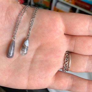 Peut inclure: Deux colliers en argent avec des pendentifs en agate en forme de larme. Les pendentifs sont gris clair avec des tourbillons blancs et bruns.
