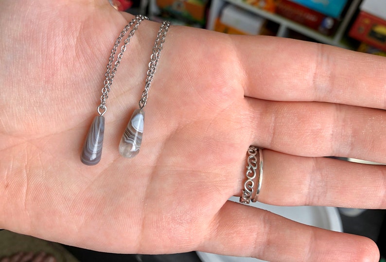 Peut inclure: Deux colliers en argent avec des pendentifs en agate en forme de larme. Les pendentifs sont gris clair avec des tourbillons blancs et bruns.