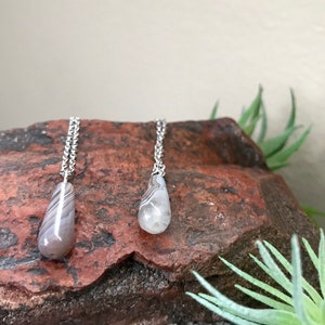 Peut inclure: Deux colliers en argent avec des pendentifs en agate en forme de goutte. Les pendentifs sont de couleur gris clair avec des bandes brunes et blanches.