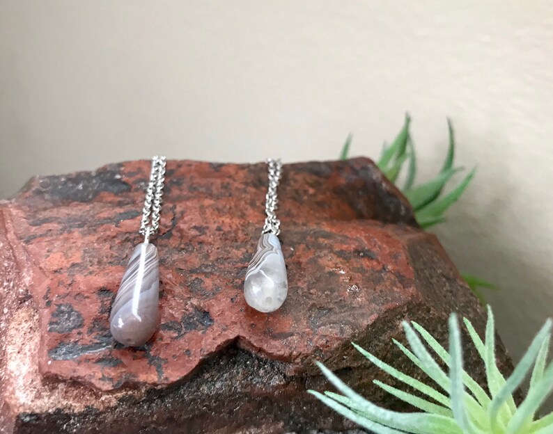 Peut inclure: Deux colliers en argent avec des pendentifs en agate en forme de goutte. Les pendentifs sont de couleur gris clair avec des bandes brunes et blanches.