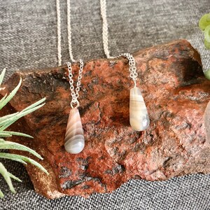 Peut inclure: Deux colliers en argent avec des pendentifs en agate en forme de larme. L'agate est un m&eacute;lange de couleurs blanches, grises et brunes. Les colliers sont pr&eacute;sent&eacute;s sur une roche rugueuse de couleur brun rouge.