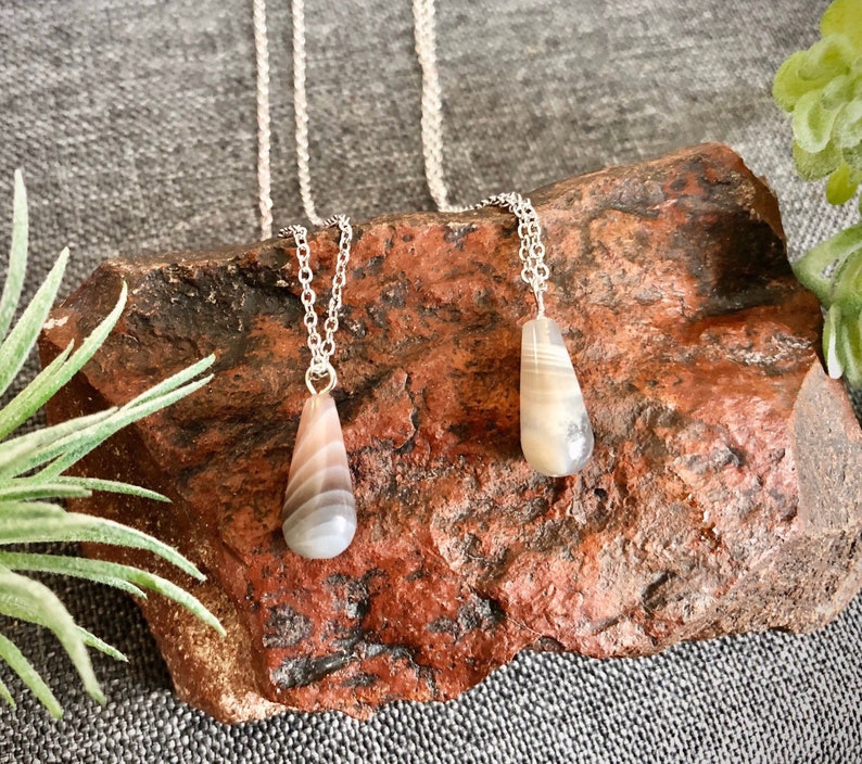 Peut inclure: Deux colliers en argent avec des pendentifs en agate en forme de larme. L'agate est un m&eacute;lange de couleurs blanches, grises et brunes. Les colliers sont pr&eacute;sent&eacute;s sur une roche rugueuse de couleur brun rouge.