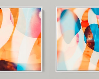 May include: Two framed abstract art pieces. Each features a vibrant mix of colors, including orange, blue, pink, and white, with flowing, organic shapes. The artwork is encased in a white frame, creating a modern aesthetic.
