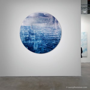 Può includere: Un dipinto circolare di un oceano blu con un cielo nuvoloso sopra. Il dipinto è esposto su una parete bianca in un ambiente di galleria.