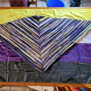 May include: A knitted triangular shawl in shades of purple, yellow, and white is draped over a purple, white, and black flag. The flag is the non-binary pride flag.