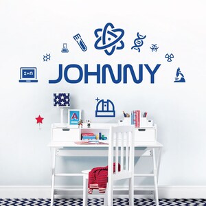 Puede incluir: Un escritorio y una silla blancos con un suéter a rayas rojas y blancas sobre la silla. La pared presenta el nombre "JOHNNY" en azul, rodeado de iconos de temática científica. También se ven un gancho en forma de estrella y una lámpara con un diseño de estrellas.