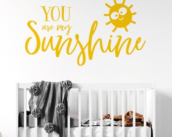 Peut inclure: Un autocollant mural jaune avec l'inscription "YOU are my Sunshine" et un soleil souriant. En dessous, un lit bébé blanc avec une couverture grise et des pompons est visible, créant un thème de chambre d'enfant joyeux.