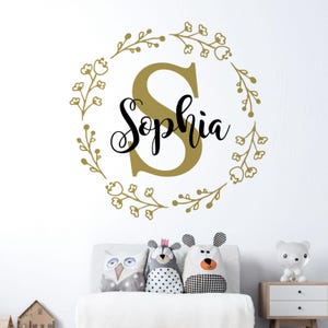 Może przedstawiać: Złota naklejka na ścianę w kształcie wieńca kwiatowego z imieniem "Sophia" w czarnym stylu pisma wewnątrz wieńca.