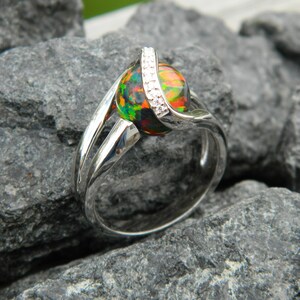 May include: A silver ring featuring a round, multi-coloured opal gemstone set in a twisted band. The opal has a black base with flashes of green, red, and orange. The band is accented with small, clear diamonds.