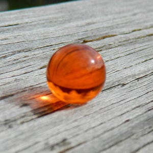 May include: A round, orange glass marble with a slight shimmer. The marble is sitting on a light brown wooden surface.