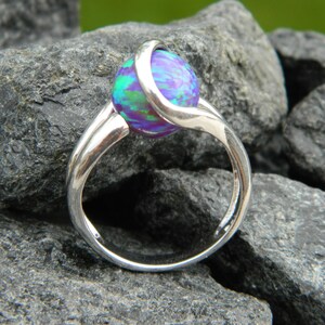 Puede incluir: Un anillo de plata con una piedra redonda de ópalo iridiscente. El anillo tiene un diseño de banda retorcida.