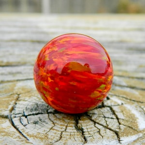 Puede incluir: Una canica de vidrio roja y naranja con un patrón de remolino. La canica es redonda y tiene un acabado brillante.