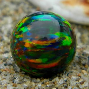 Puede incluir: Una piedra preciosa de ópalo negro redonda e iridiscente con destellos verdes, azules y naranjas. El ópalo es liso y pulido.