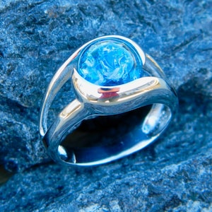 May include: A silver ring with a large, round, blue gemstone set in a bezel setting. The ring has a unique, modern design with a curved band.