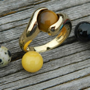 May include: A gold ring featuring a large, amber-coloured gemstone. Several colourful gemstone spheres, including black, red, blue, and turquoise, are arranged around the ring on a weathered wooden surface. Other spheres are also visible.