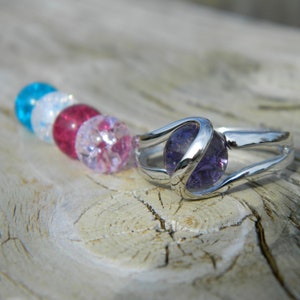 May include: A silver ring with a purple gemstone set in a swirl design. The ring is surrounded by a row of colorful beads.