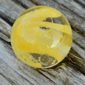 May include: A yellow glass marble with white and gray swirls inside. The marble is sitting on a wooden surface.