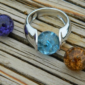 May include: A silver ring with a light blue glass stone set in the centre. The ring is surrounded by five small glass marbles in various colours, including red, purple, brown, and white.