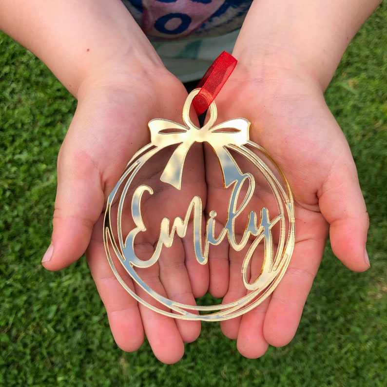 May include: Gold-colored Christmas ornament with the name "Emily" cut out in a decorative script font. The ornament is held in the hands of a person.