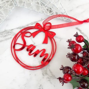 May include: Red acrylic Christmas ornament with the name "Sam" cut out in a circle with a red bow. The ornament has a red ribbon for hanging.