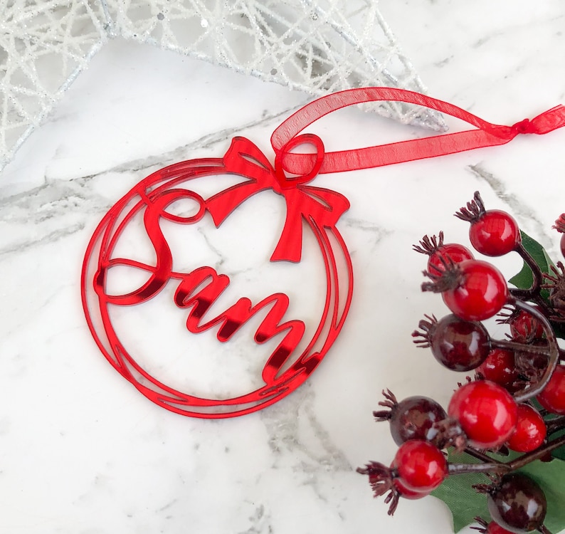 May include: Red acrylic Christmas ornament with the name "Sam" cut out in a circle with a red bow. The ornament has a red ribbon for hanging.